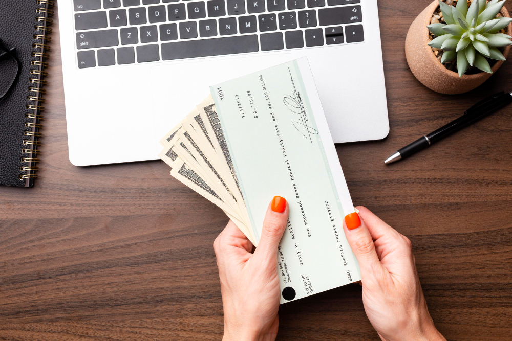 A person holding cash and a checkbook for payday loans next to a laptop on a wooden desk.