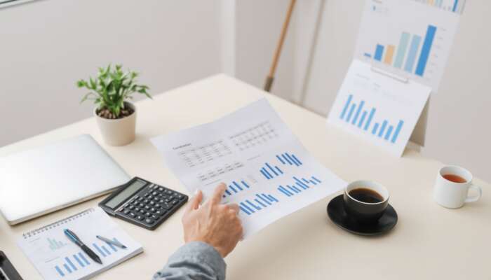 A person reviews printed charts with blue bar graphs at a desk, analyzing 2025 payday loan trends. A calculator, notebook, laptop, coffee cup, and plant are on the desk. More charts on a board appear in the background.